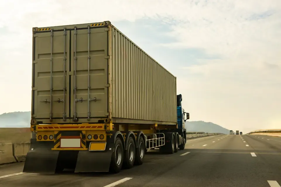 Blue semi truck hauling a beige container trailer on a highway under a bright, partly cloudy sky